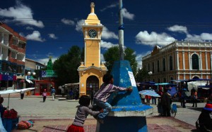 The Uyuni town square in the sunshine in December, 2005. Features children playing by a small monument and a beautiful colonial-style building in the background.